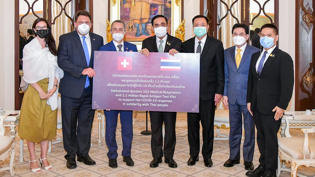 Verschiedene Personen stehen in einem Raum vor einem Schild, welches die Hilfslieferung der Schweiz beschreibt.