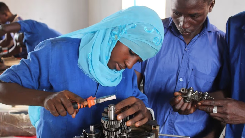 Mädchen mit Kopftuch arbeitet in einer Mechanikerlehrwerkstätte, zwei Jungs schauen zu.