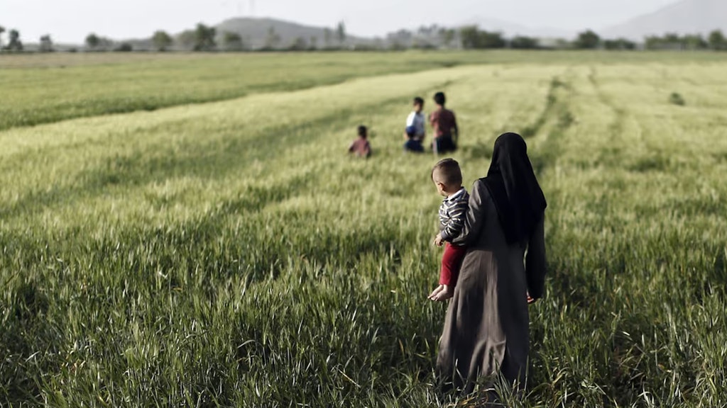 Eine Frau steht mit einem Kleinkind auf dem Arm in einem Feld mit hohem Gras und schaut Kindern in der Ferne beim Spielen zu.