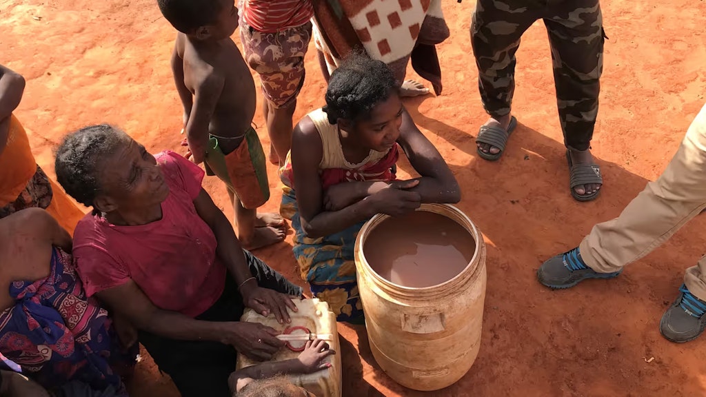 Das Bild zeigt Frauen, Kinder und einen bewaffneten Mann, die sich um einen Kessel mit dreckigem Wasser scharen.  