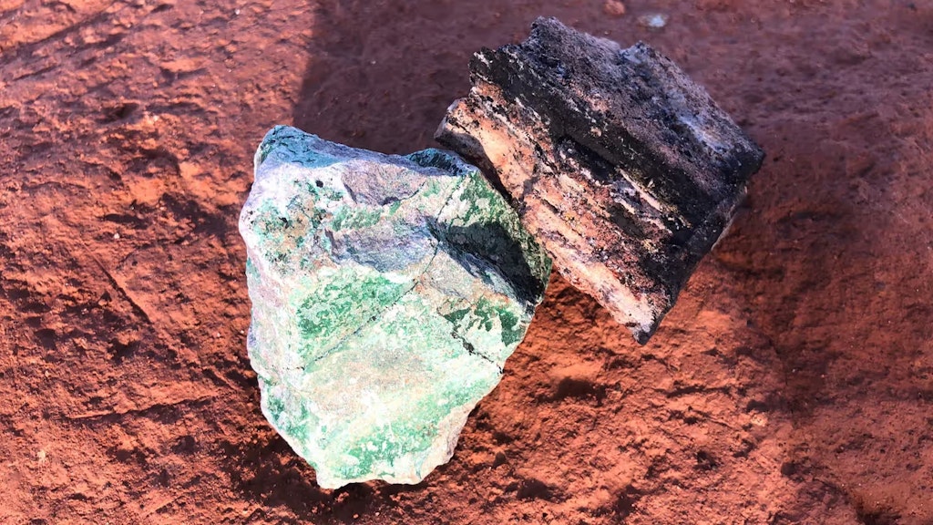 Ein Stück schwarzes Kobalt und ein Stück grünes Kupfer liegen auf dem ockerfarbenen Boden, um den Abbau im Kupfergürtel der Demokratischen Republik Kongo zu veranschaulichen.