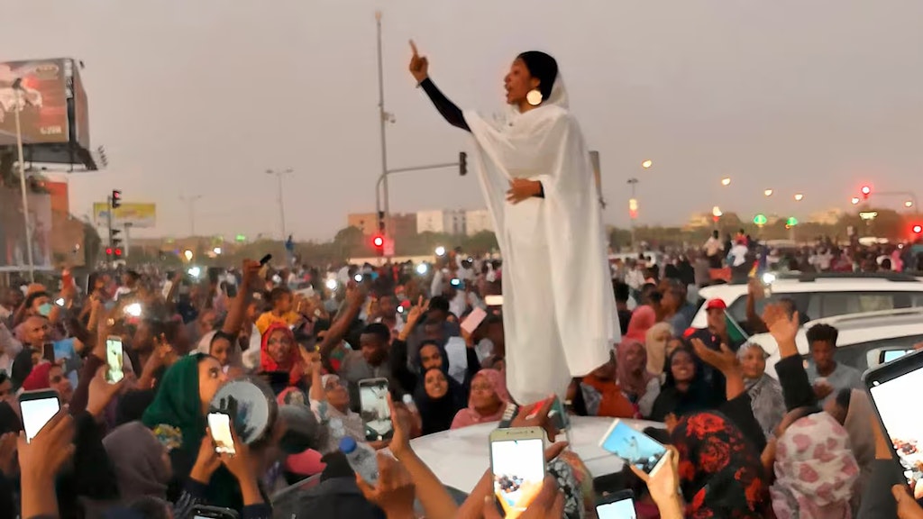 Umringt von einer Menschenmenge spricht eine Frau, die auf der Motorhaube eines Autos steht. Sie wird mit zahlreichen Handys gefilmt.
