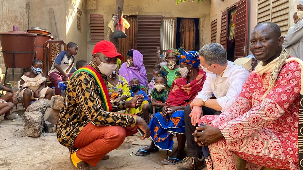 Aussenminister Cassis sitzt vor einem Haus im Kreis von afrikanischen Gesprächspartnern, Frauen, Männern und Kindern.