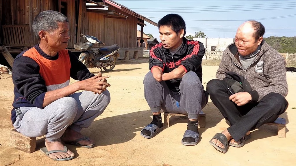 Drei laotische Streumunitionsopfer sitzen auf dem Boden vor einem Bauernhaus.