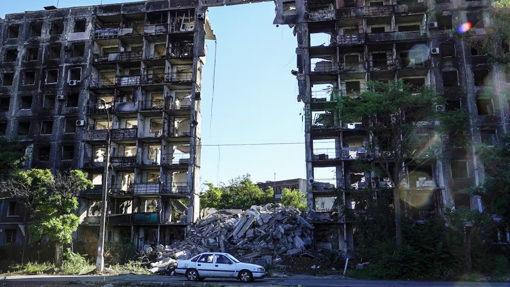 Ein weisses Auto steht vor einem durch Beschuss zerstörtem zivilen Gebäude, durch das man den blauen Himmel sieht.