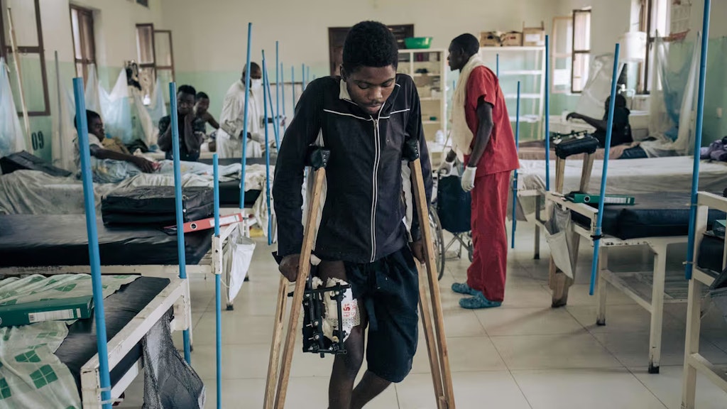 Ein junger afrikanischer Mann mit einer Schusswunde am Bein geht in einem Krankenhaus mühsam auf Krücken.