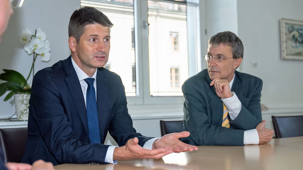 Gabriel Lüchinger sitzt an einem Tisch und spricht. Franz Perrez sitzt rechts neben ihm und hört ihm zu.
