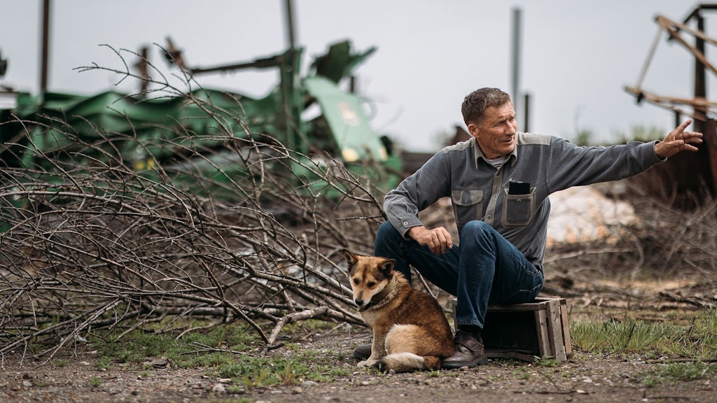 Ein ukrainischer Bauer sitzt auf einer Holzkiste inmitten von Minenräumungsgeräten. Vor ihm sitzt sein Hund.