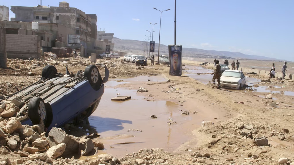 Ein Auto liegt auf dem Dach in Schlamm und Mauerresten. Menschen suchen im Hintergrund nach Opfern der Überschwemmung.