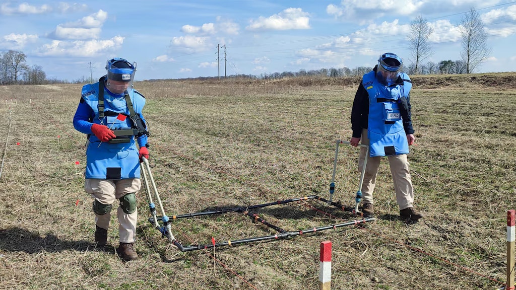 Zwei Männer in Schutzkleidung arbeiten an der Minenräumung auf einem Feld, auf dem sich wahrscheinlich Munition befindet.