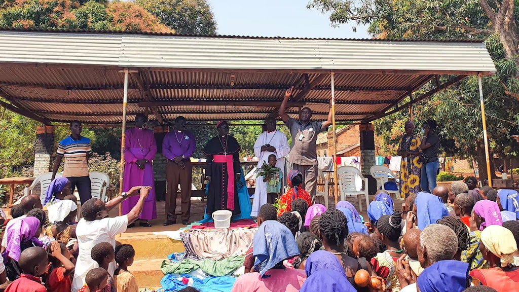 Auf einem Podium stehen fünf Männer, die aufgrund ihrer Kleidung als Vertreter verschiedener Kirchen erkennbar sind. Einer davon spricht zum Publikum, das im Vordergrund des Bildes zu sehen ist.