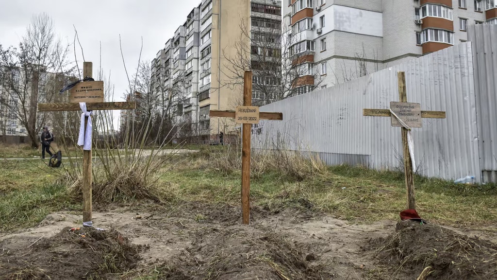 Drei Gräber mit Holzkreuzen und Namen sind im Vordergrund zu sehen. Im Hintergrund stehen grosse Häuser.