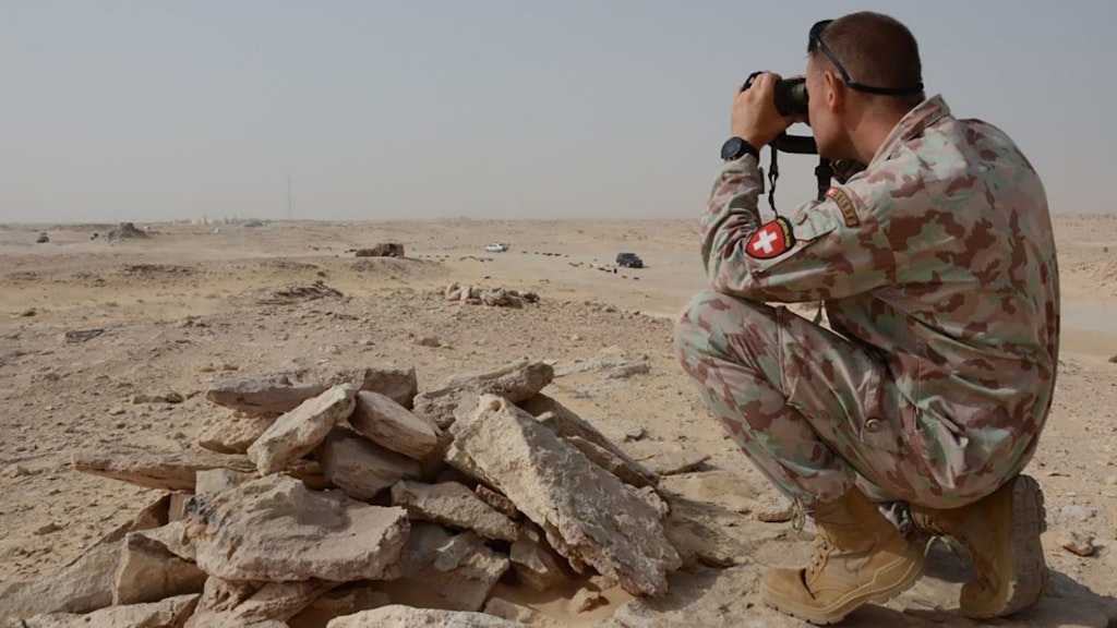 Ein Soldat der Schweizer Armee kniet im Sand und schaut mit dem Fernglas in die Wüste.