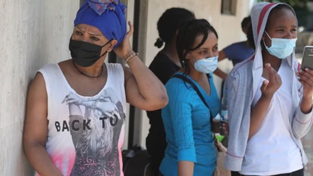 Drei Frauen mit Masken warten vor einem Gebäude. Einer von ihnen hält ein Telefon in der Hand.