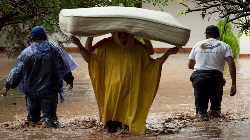 Zwei Personen tragen eine Matratze durch das Hochwasser. Zwei Mitglieder der Hilfskräfte begleiten sie dabei.