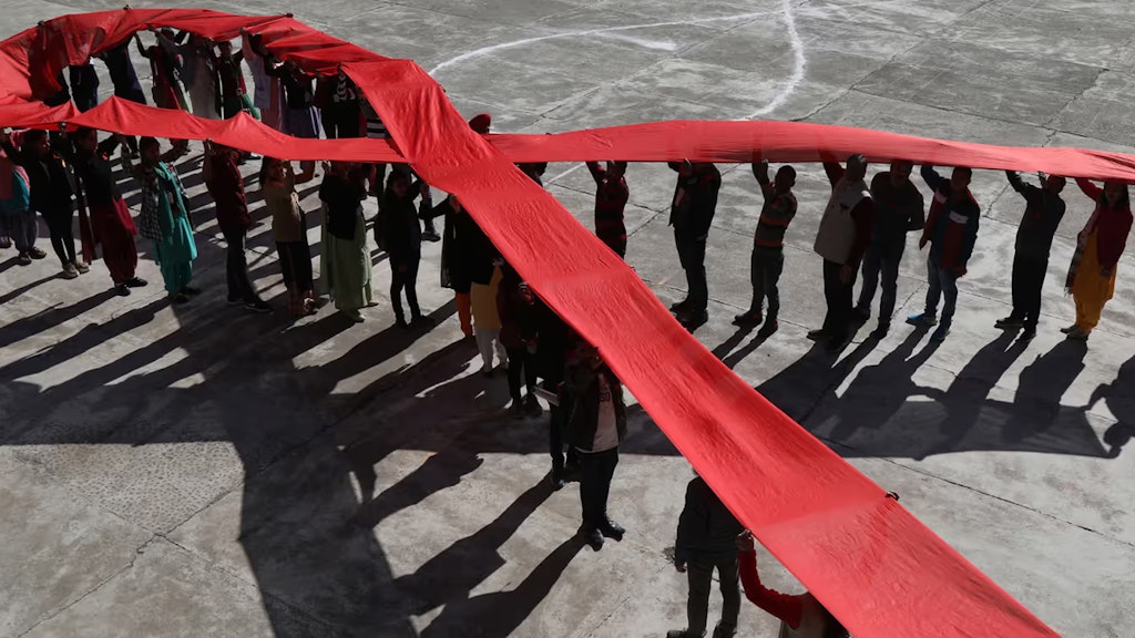 Viele Personen heben ein langes rotes Band in Form einer Schlaufe über ihren Köpfen. Sie ist weltweit ein Symbol für den Kampf gegen Aids.