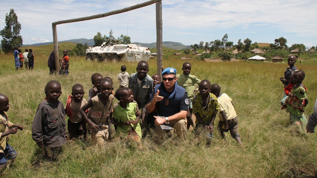 Polizist in der DRC. Draussen in einem grünen Feld mit einer Gruppe Kinder. Im Hintergrund ist ein weisses gepanzertes Fahrzeug der UN zu sehen.
