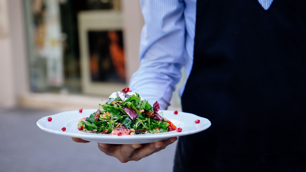 A man in a striped shirt wearing an apron serving a colourful salad. 