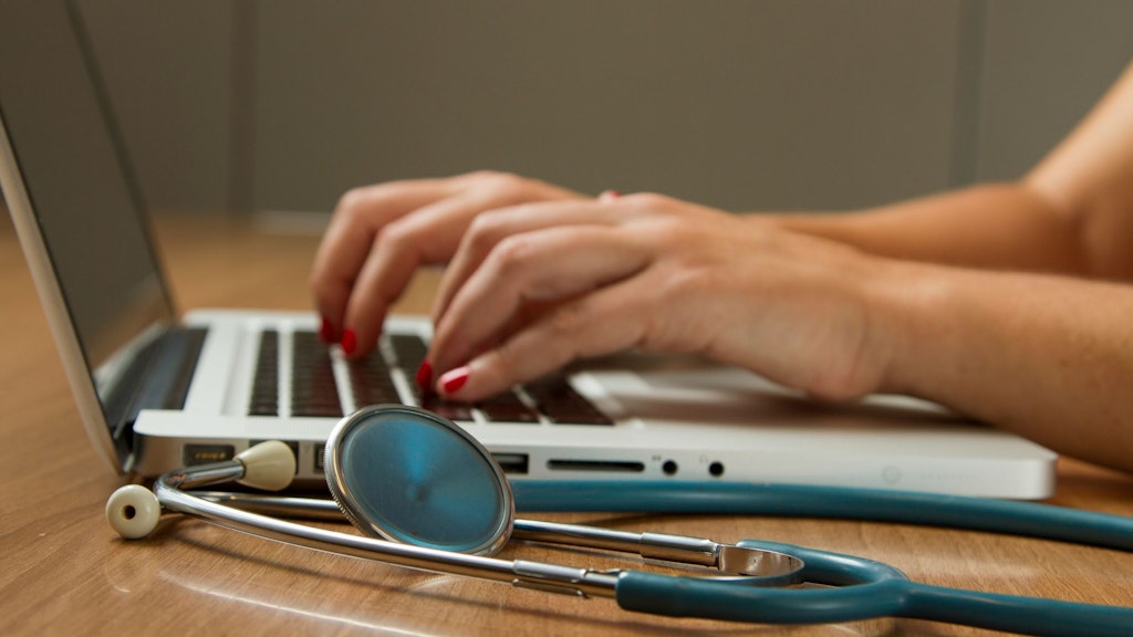 A stethoscope lies on a table in front of a laptop on which a woman is typing.