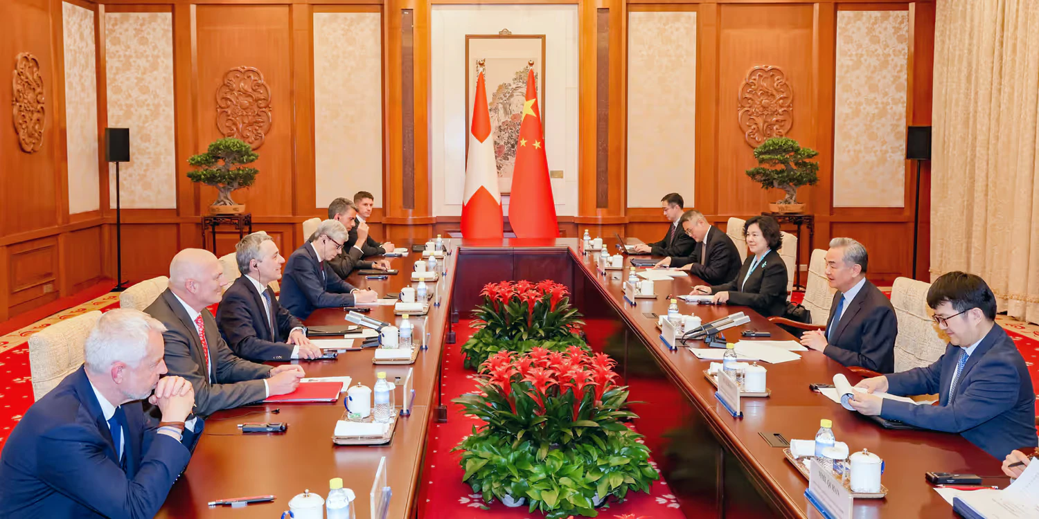 Die Delegationen der Schweiz und Chinas sitzen an einem langen Holztisch. Im Hintergrund sind die Flaggen der Schweiz und Chinas zu sehen.