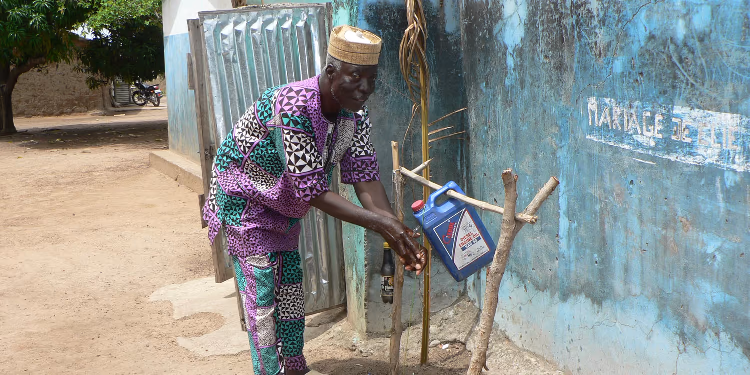 Ein Mann steht in Benin vor einer Hütte und wäscht sich an einer improvisierten Sanitäranlage gründlich die Hände.