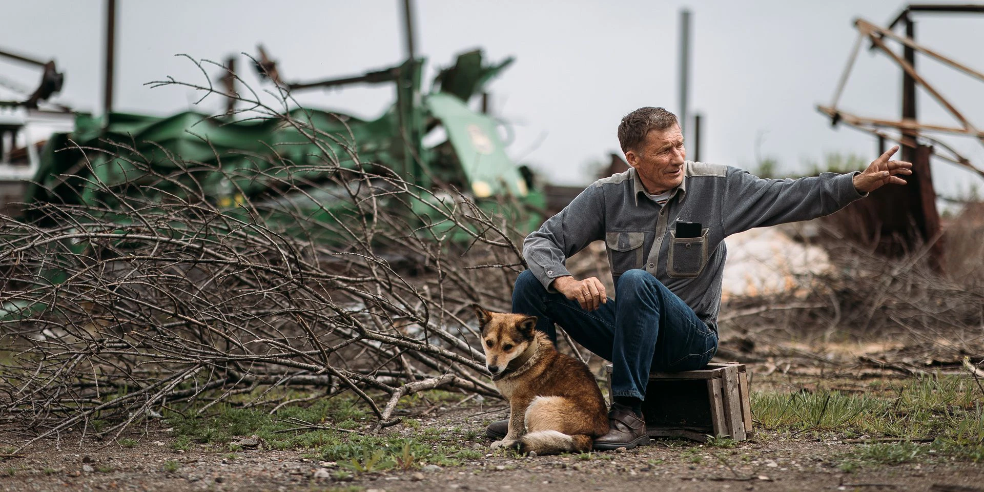 Ein ukrainischer Bauer sitzt auf einer Holzkiste inmitten von Minenräumungsgeräten. Vor ihm sitzt sein Hund.