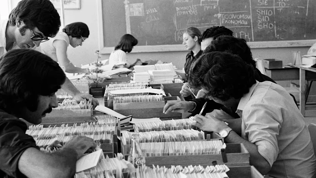 Das Bild, schwarz-weiss, zeigt einen Büroraum im Jahr 1974. Die Tische sind vollgestellt mit Karteikästen inkl. Karteikarten. Acht Personen arbeiten konzentriert daran.