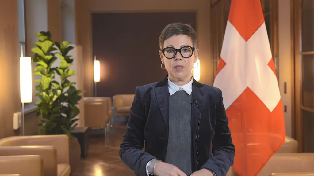 EDA-Staatssekretärin Krystyna Marty Lang steht in einem getäferten Raum und spricht in eine Kamera. Im Hintergrund ist die Flagge der Schweiz zu sehen.