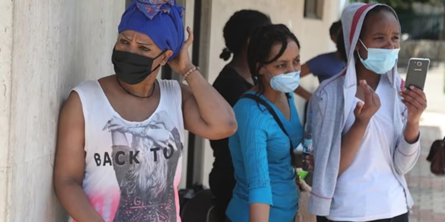 Drei Frauen mit Masken warten vor einem Gebäude. Einer von ihnen hält ein Telefon in der Hand.
