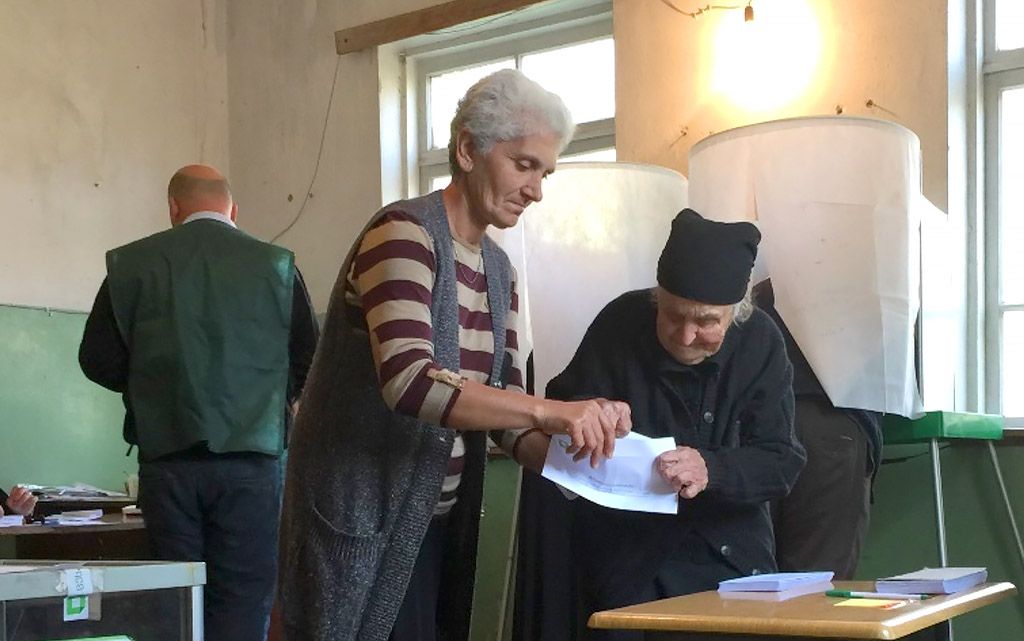 Zwei Frauen stehen bei den Kommunalwahlen in Georgien in einem Wahllokal vor einer Wahlurne. Die eine hilft der anderen beim Falten eines Stimmzettels.