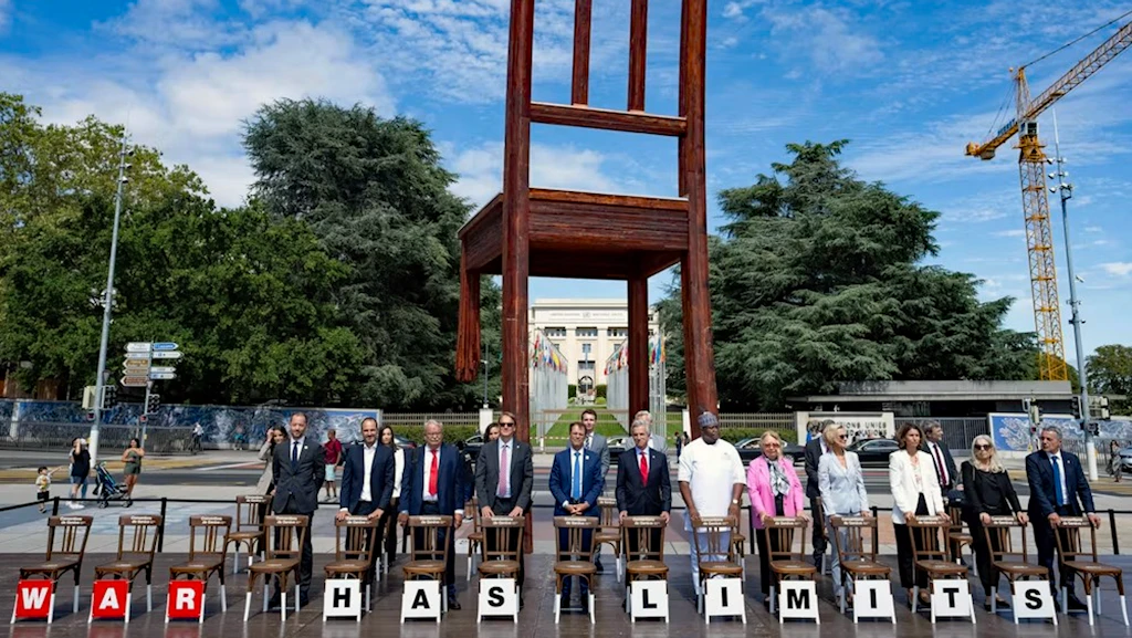 Personengruppe steht hinter leeren Stühlen mit der Aufschrift «WAR HAS LIMITS» vor dem «Broken Chair» und dem UNO-Sitz in Genf.
