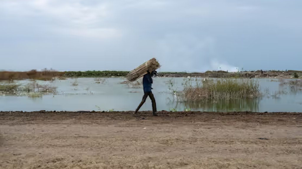 Ein Mann aus dem Südsudan trägt Holz auf seinen Schultern. Er geht an überschwemmten Flächen vorbei.