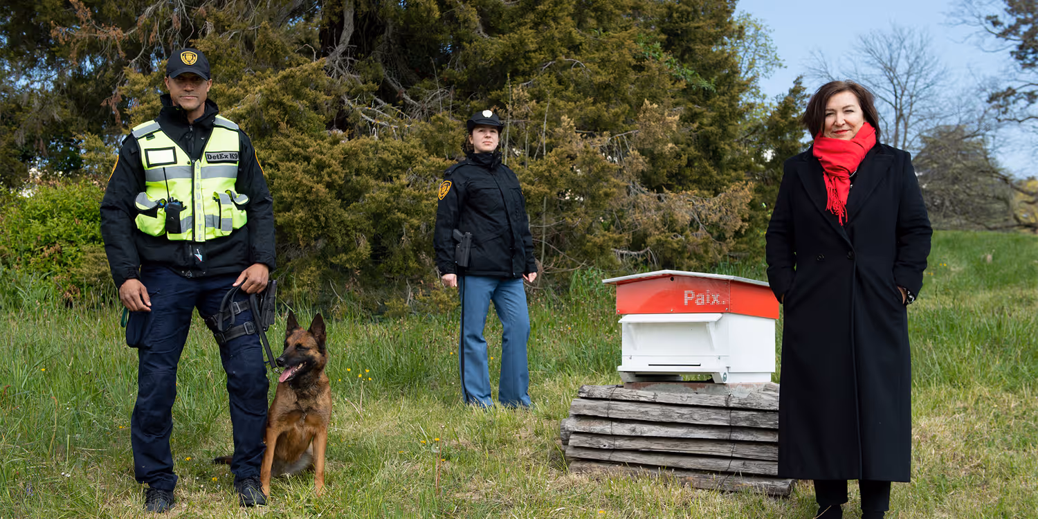 Auf dem Rasen des Gebäudes der Vereinten Nationen steht Anne-Lise Favre Pilet neben zwei UN-Agenten und einem Bombenspürhund. 