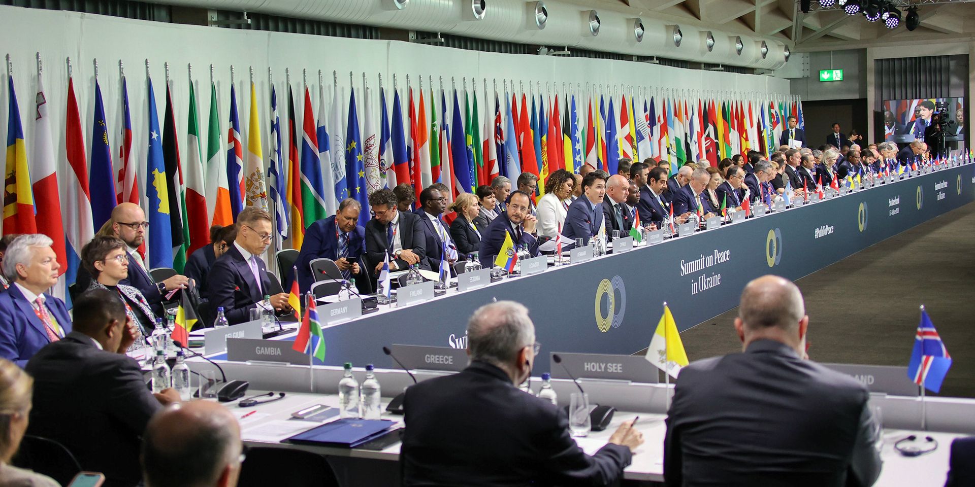 Blick in den Plenarsaal der hochrangigen Konferenz zum Frieden in der Ukraine. Im Hintergrund sind die Flaggen aller teilnehmenden Staaten und Organisationen zu sehen.