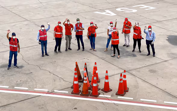 Blick aus dem Flugzeug auf eine Gruppe von Botschaftsmitarbeitern in roten Jacken, die auf dem Flugfeld steht und winkt. 