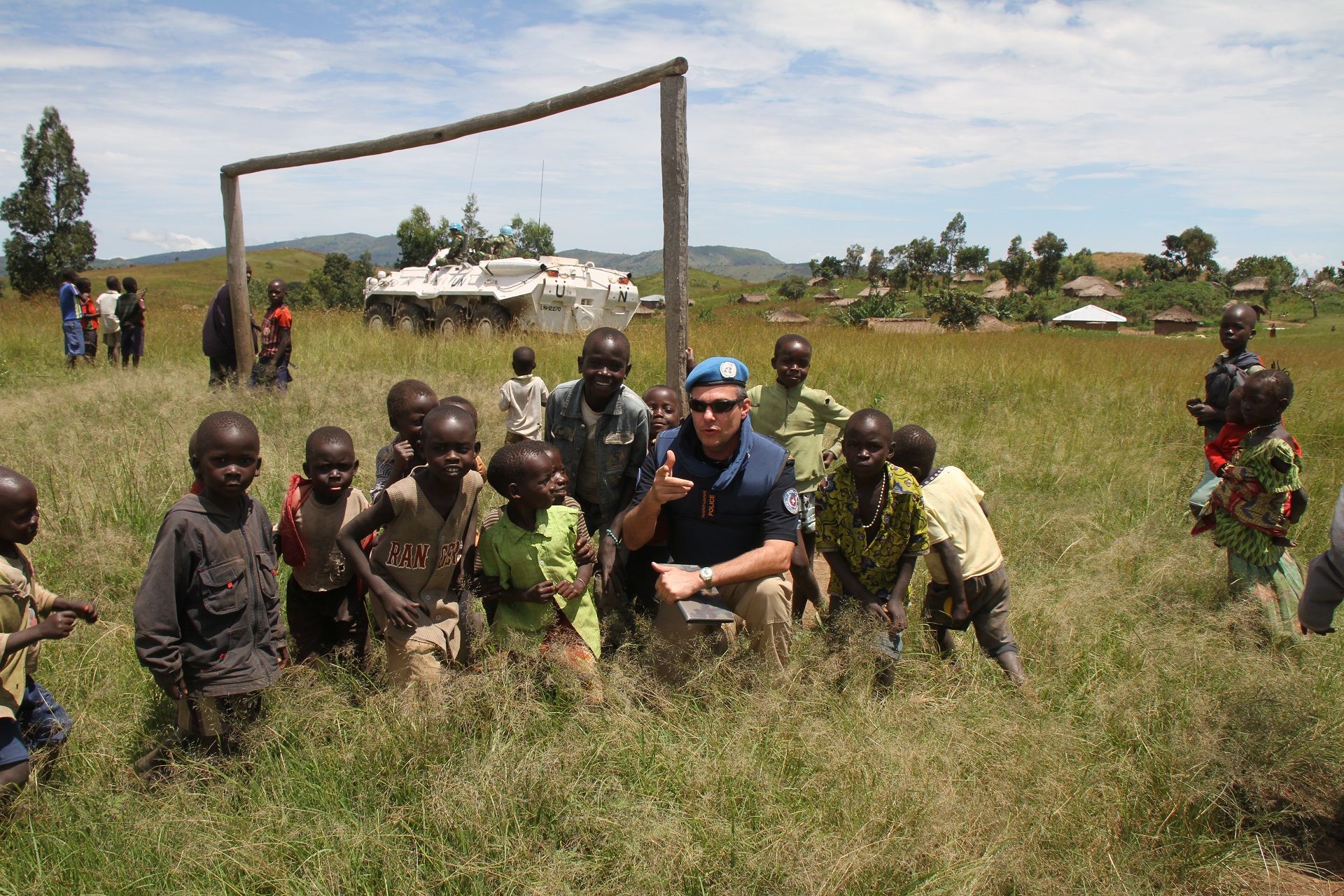 Polizist in der DRC. Draussen in einem grünen Feld mit einer Gruppe Kinder. Im Hintergrund ist ein weisses gepanzertes Fahrzeug der UN zu sehen.