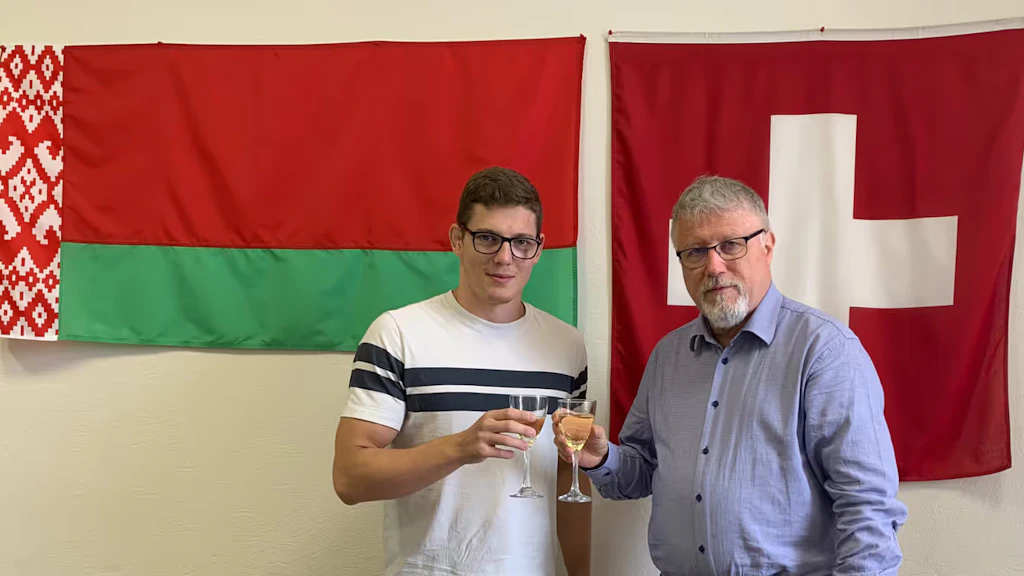 Two men stand in front of a wall with the flags of Belarus and Switzerland. 