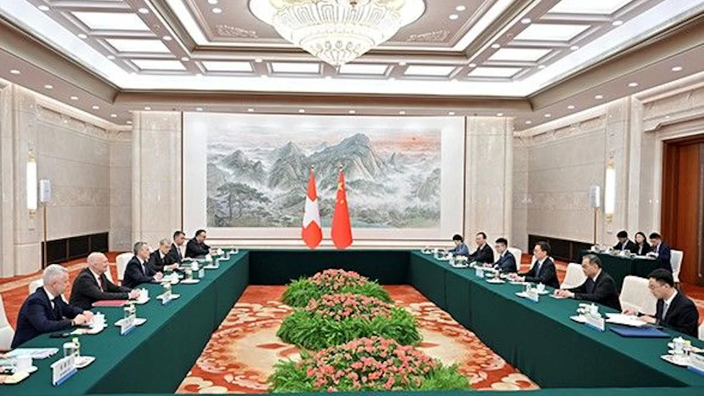 In a hall, the flags of Switzerland and China can be seen in the background. To the right and left of the room, delegations from the two countries are seated at a table.
