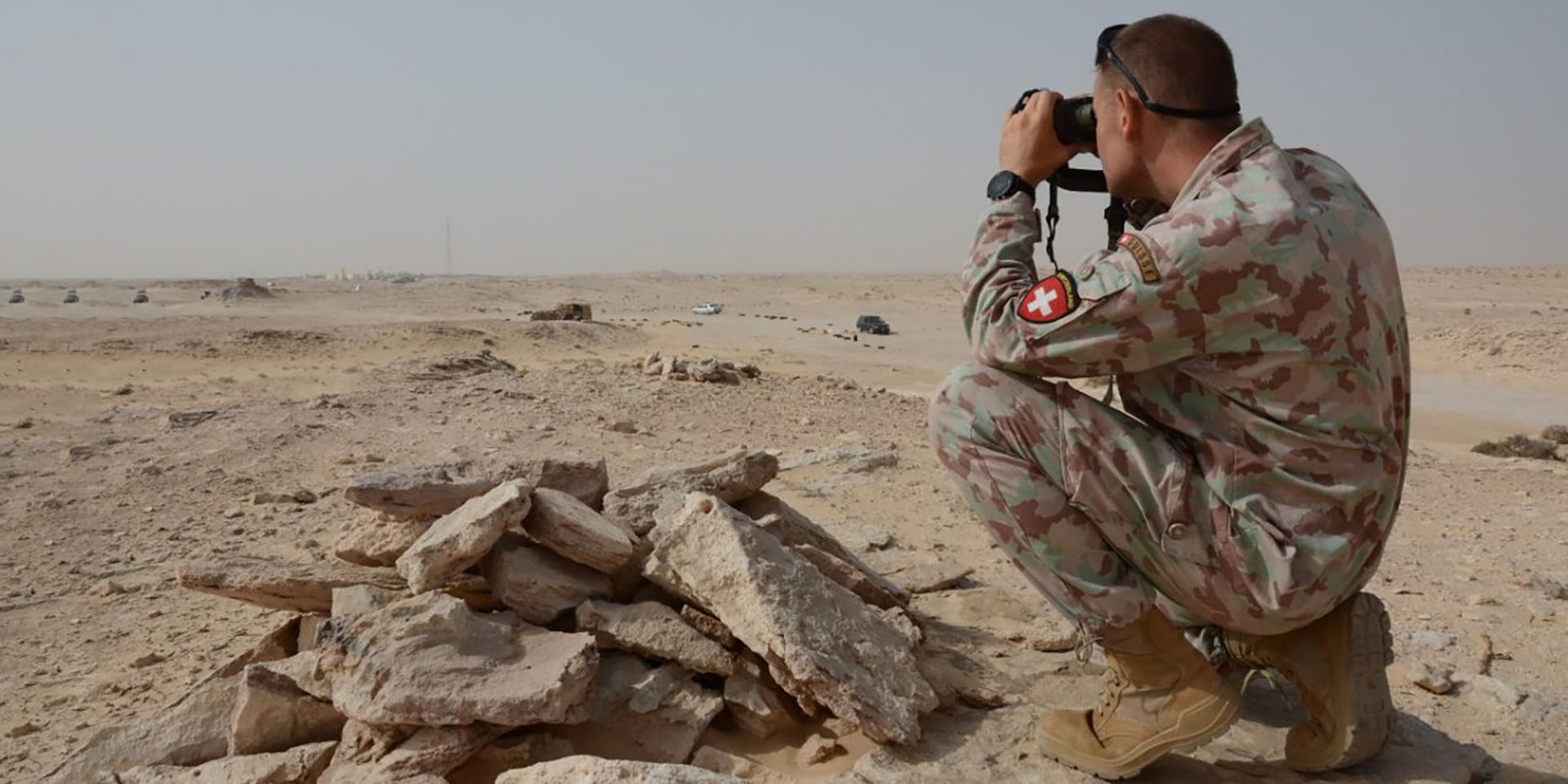 A soldier of the Swiss army kneels in the sand and looks with the binoculars into the desert.