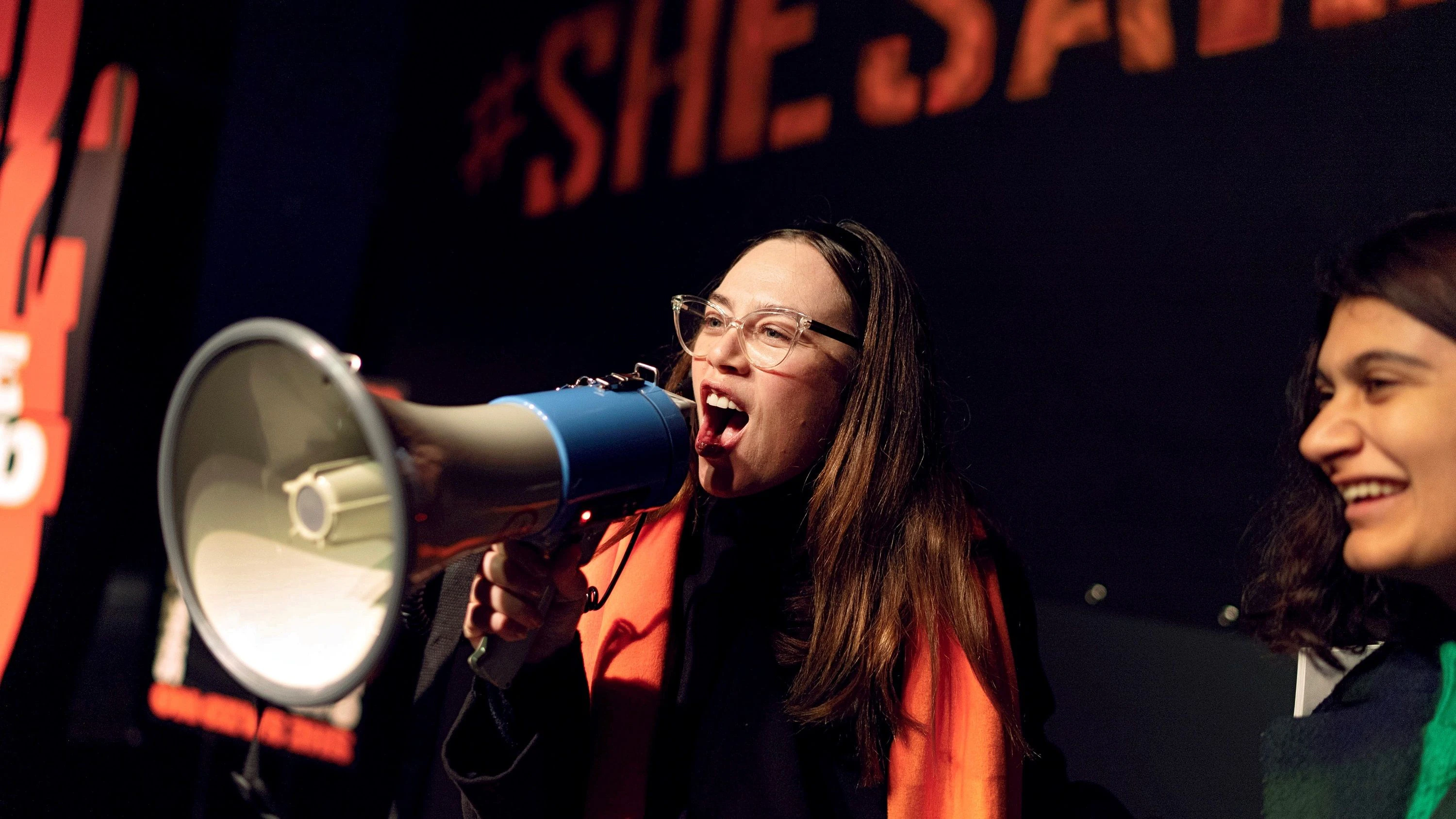 A woman wearing an orange scarf delivering a powerful message through a megaphone.