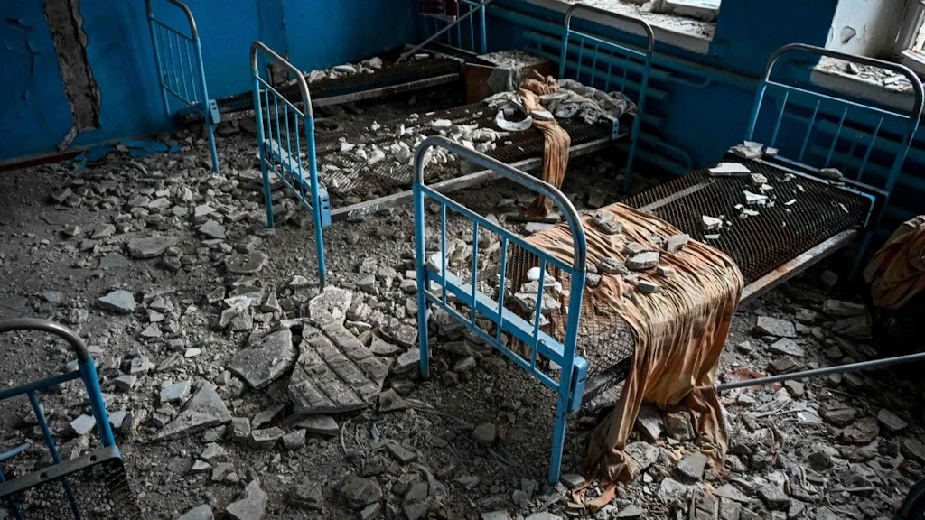  Four bedframes in a room with damaged walls and windows. The floor is littered with rubble.
