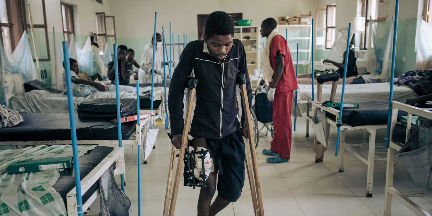 A young African man with a gunshot wound to the leg walks with difficulty on crutches in a hospital.