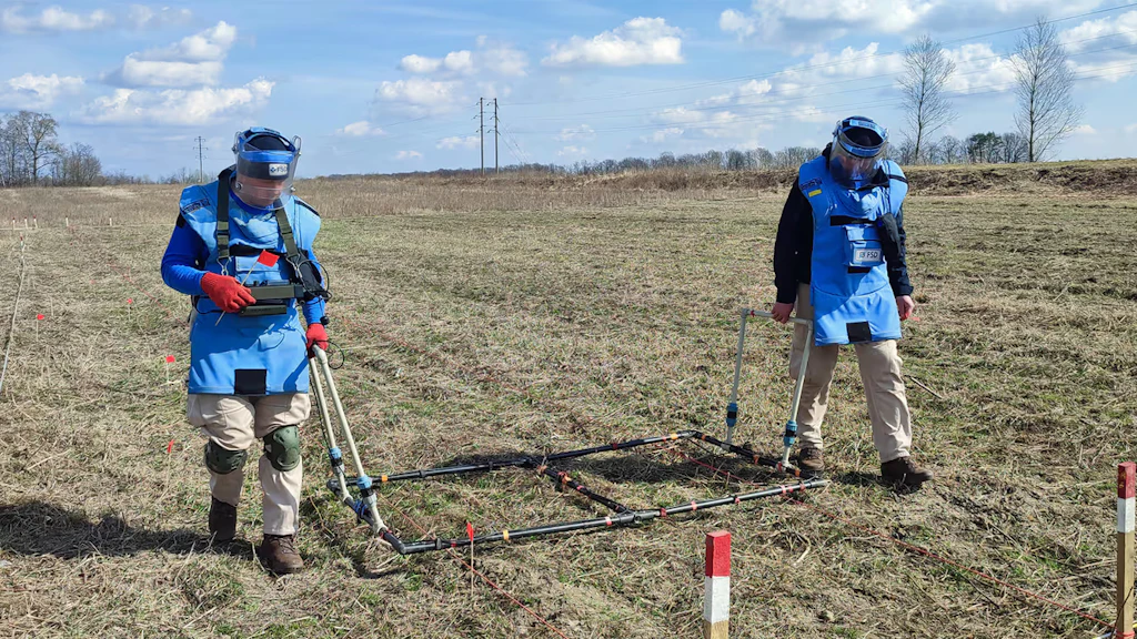 Two men in protective clothing are working on clearing a field of suspected ammunition.