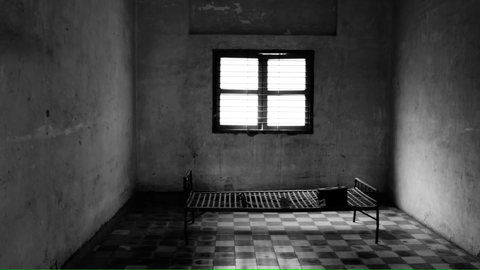 A cell with a small window and a dilapidated bed with no mattress in the middle.