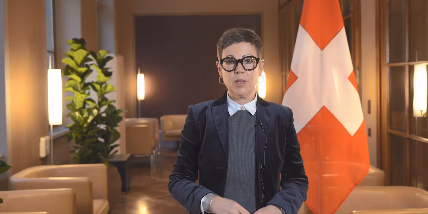 FDFA State Secretary Krystyna Marty Lang stands in a wood-panelled room and speaks into a camera. The Swiss flag can be seen in the background.