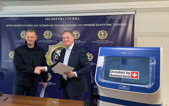 Ambassador Claude Wild shakes hands with a representative of the Ukrainian authorities. In the foreground are DNA analysis machines with the sticker "Donated by Switzerland".