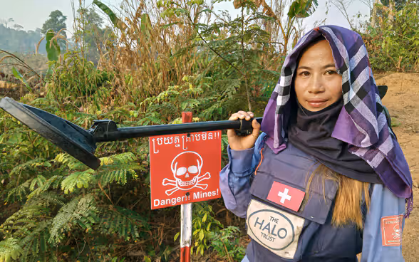 A woman deminer from the HALO Trust wearing an outfit marked with a Swiss cross standing next to a minefield.