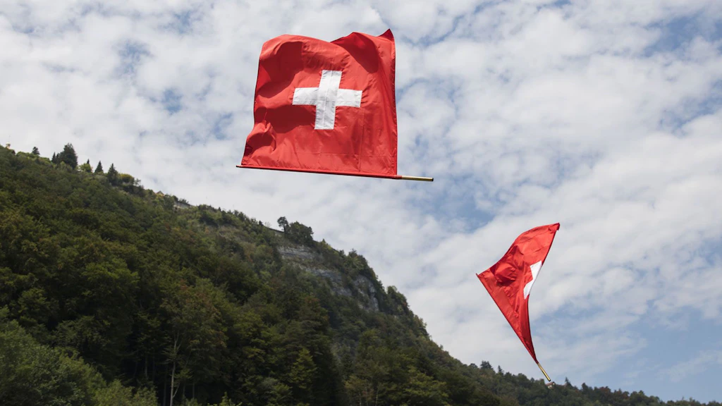 Two Swiss flags fly in the air near a mountain. 