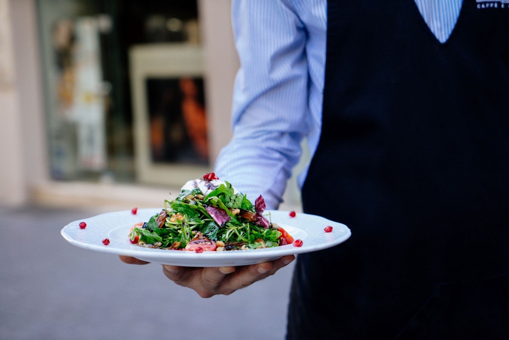 A man in a striped shirt wearing an apron serving a colourful salad. 