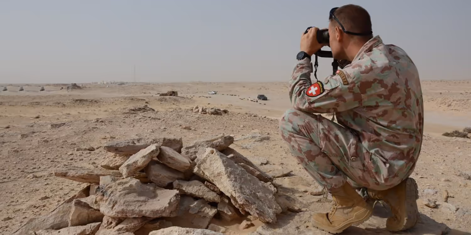 A soldier of the Swiss army kneels in the sand and looks with the binoculars into the desert.